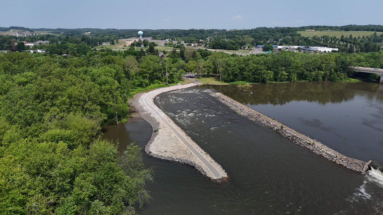 An aerial view of a river with a rocky shoreline extending into the water. Lush green trees line the banks, and a water tower is visible in the background along with a small town.