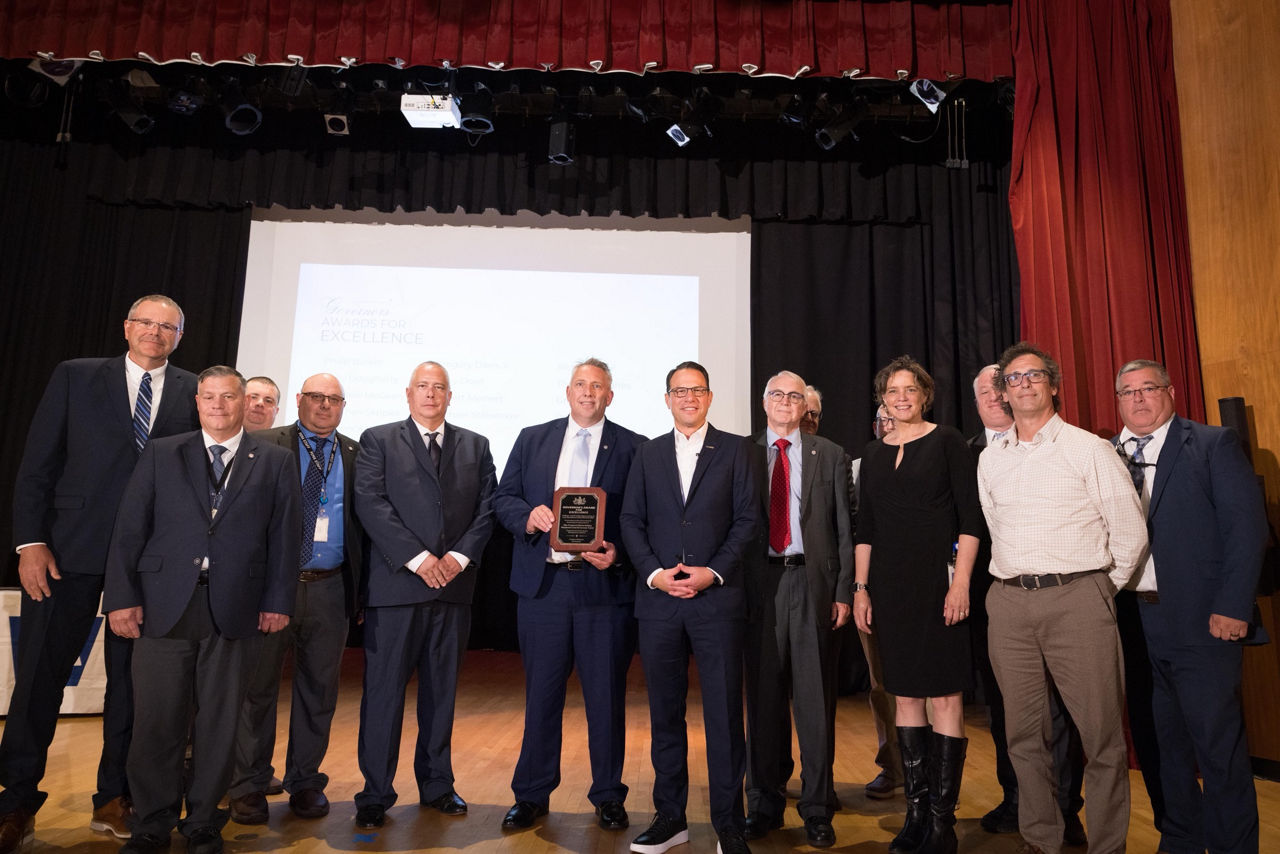 Thirteen members of PEMA staff stand with Governor Josh Shapiro on stage during an award ceremony.