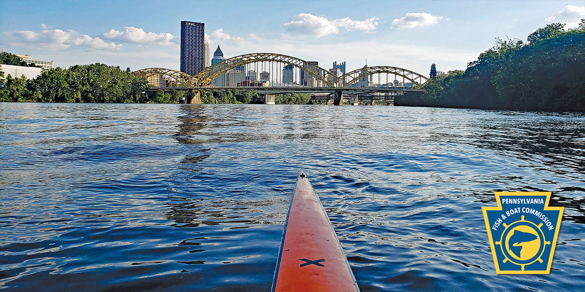 Tip of a long river kayak overlooking a beautiful, scenic view of the Pittsburgh skyline with the PFBC keystone logo in bottom right corner.