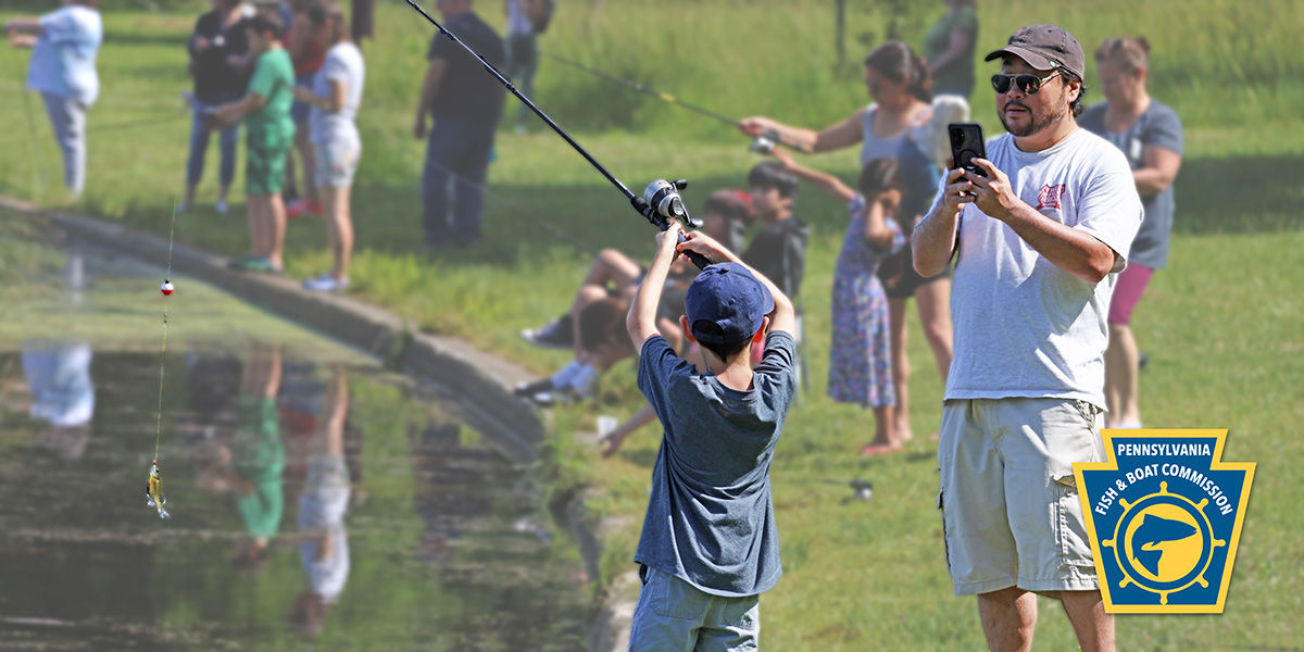 A father takes a photo with his cell phone of a young child holding up a fish he caught with his fishing rod.