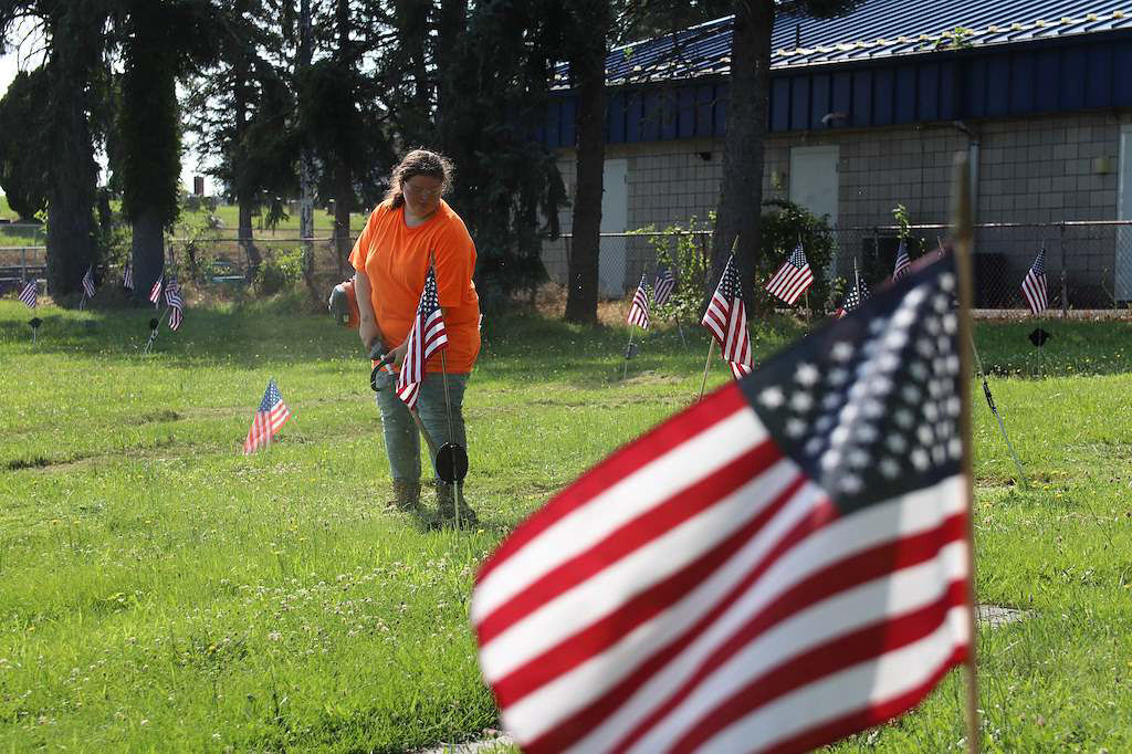 A MY Work student in an orange shirt tends to grass in a field adorned with American flags. The sunny setting conveys respect and remembrance.