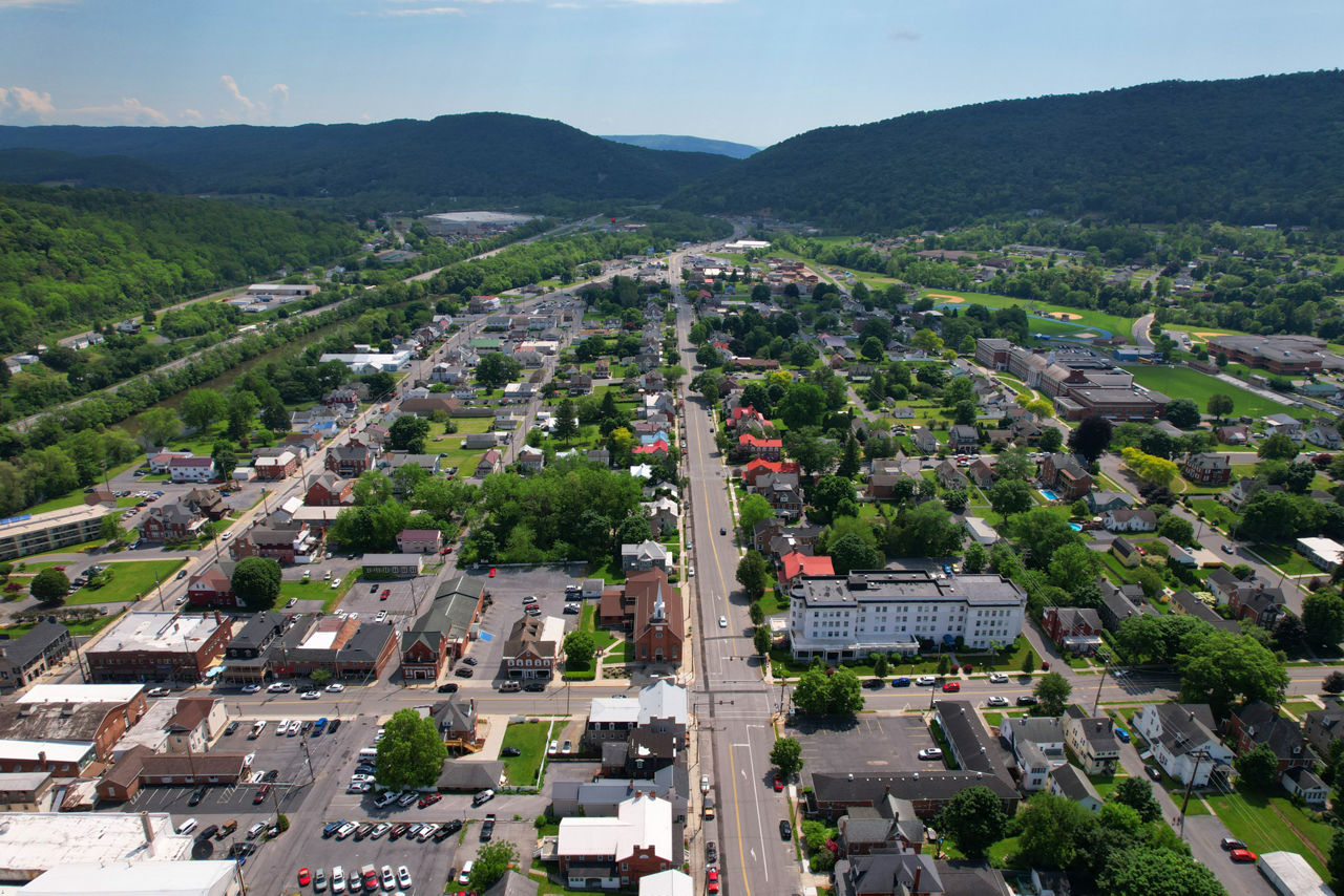 An aerial image of Bedford, PA. Buildings, roads and foliage extend into the distance, surrounded by forest-covered mountains and a blue sky.