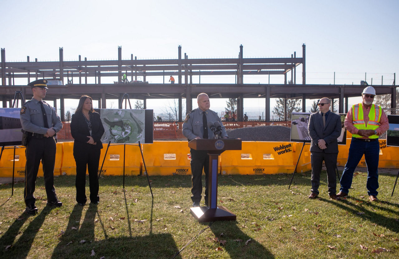 State Police Lieutenant Colonel Bivens speaks at a podium in front of a construction site with a steel beam framed building. Four other people stand alongside him. Aerial photos and architectural images are displayed on easels.