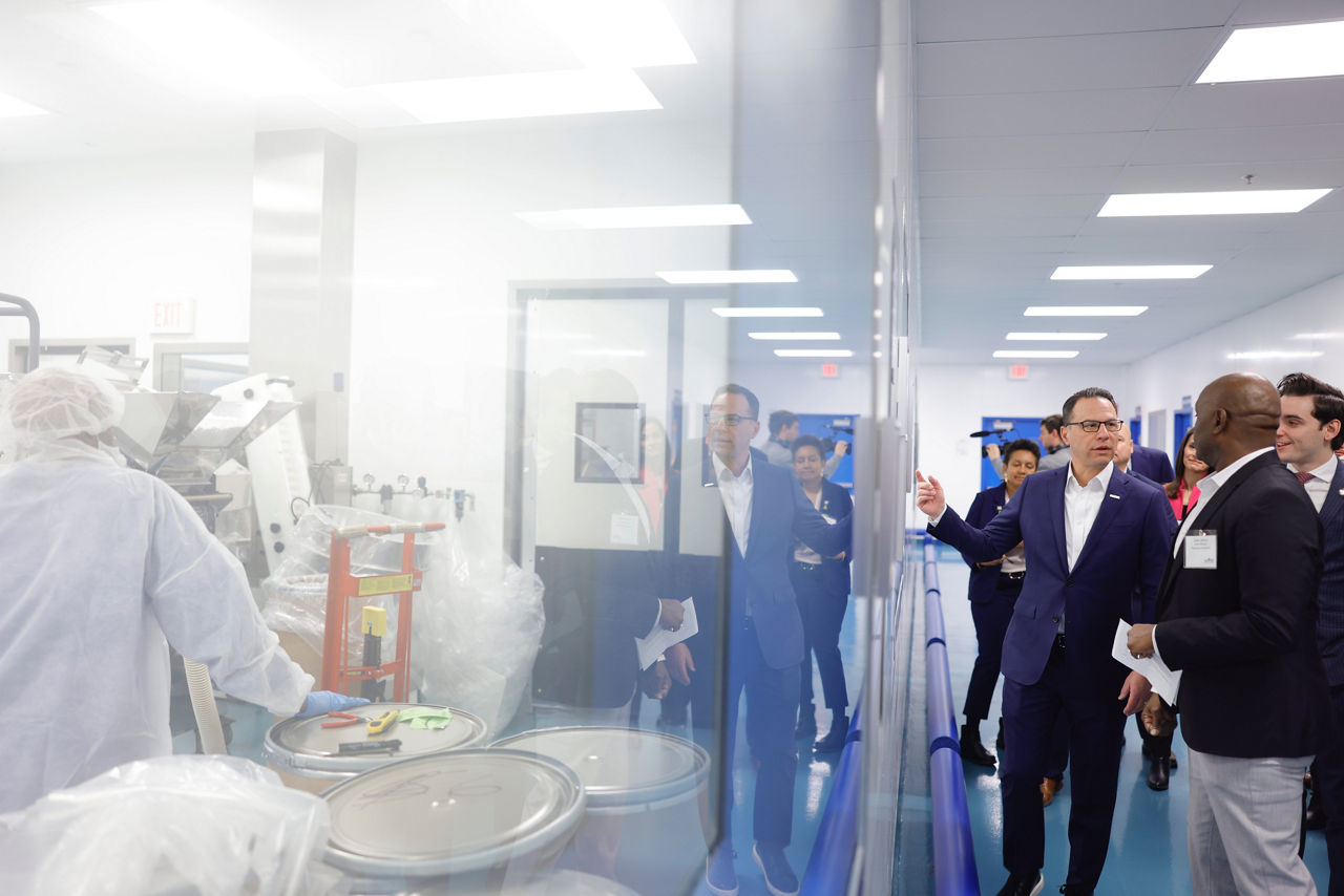 Gov. Josh Shapiro and executives tour a biopharma facility, viewing lab workers working through a large glass window.
