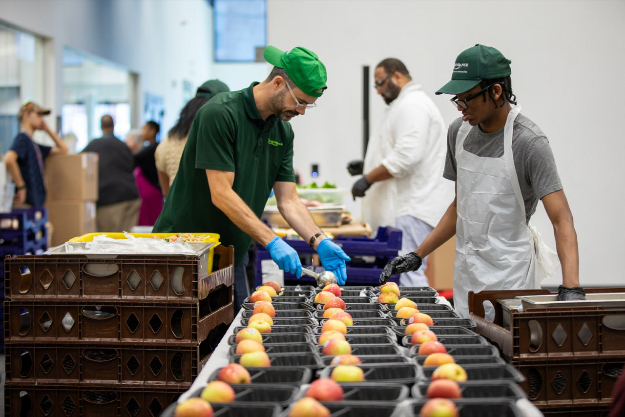 Two adults help place apples into packaged food trays