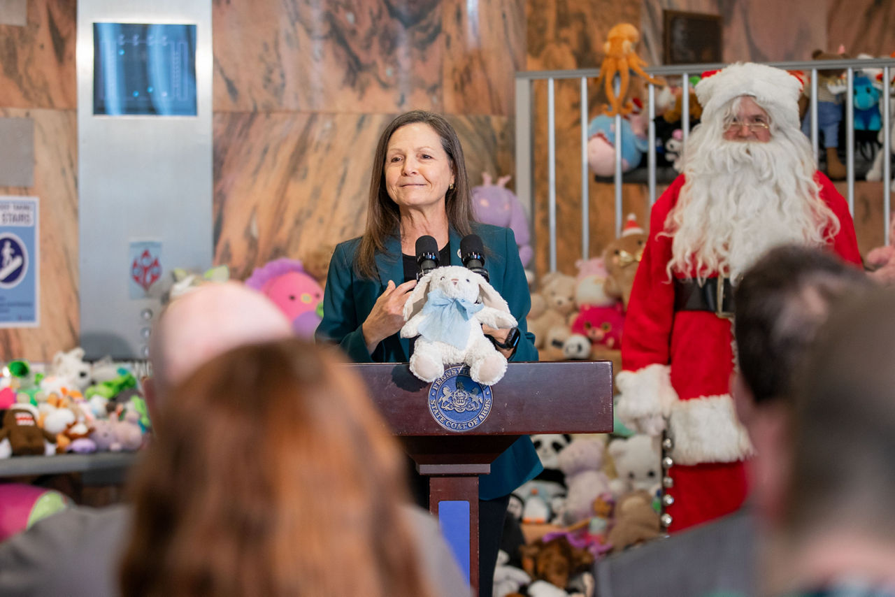 L&I Secretary Nancy A. Walker addresses audience at podium in front of Santa Claus and hundreds of safety-inspected stuffed toys.