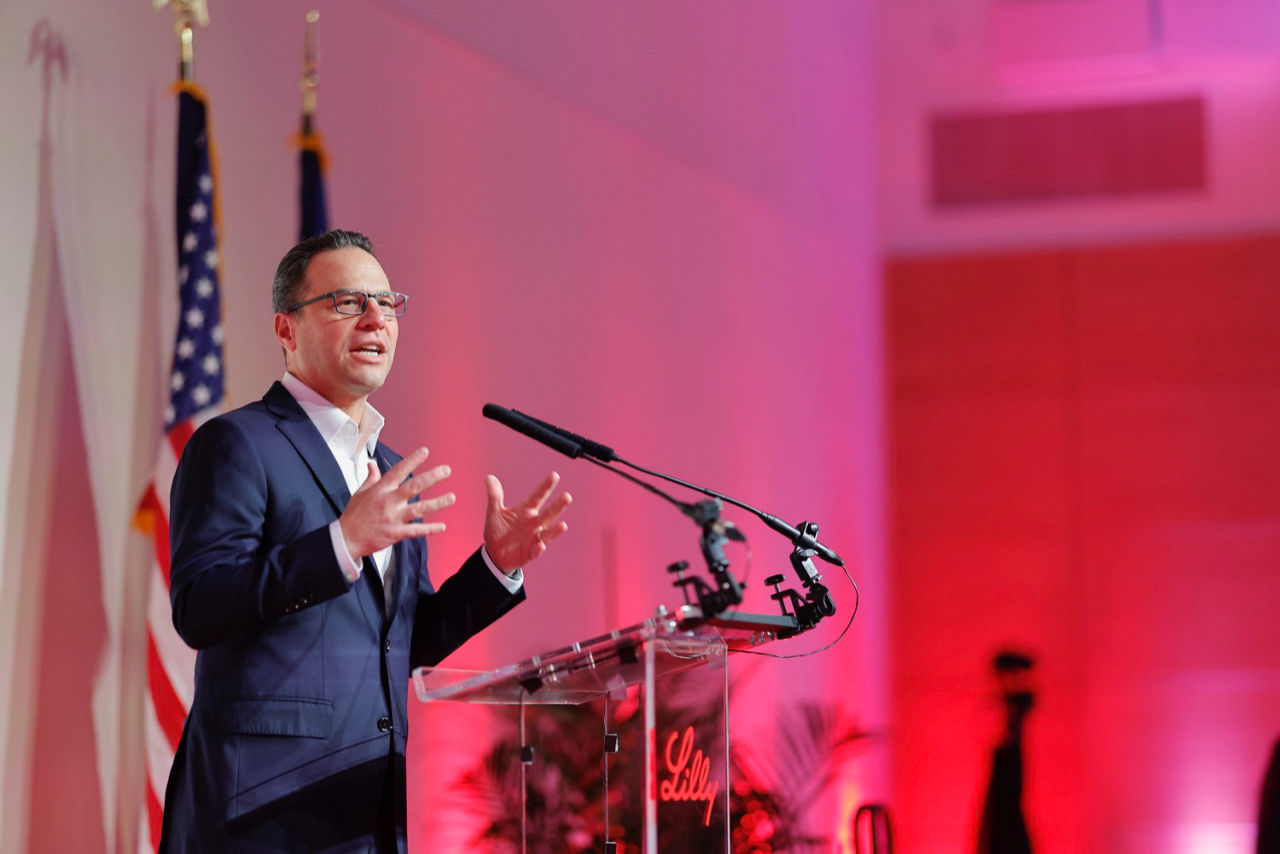 Governor Josh Shapiro speaks at a podium against a backdrop of US flags and red lighting.