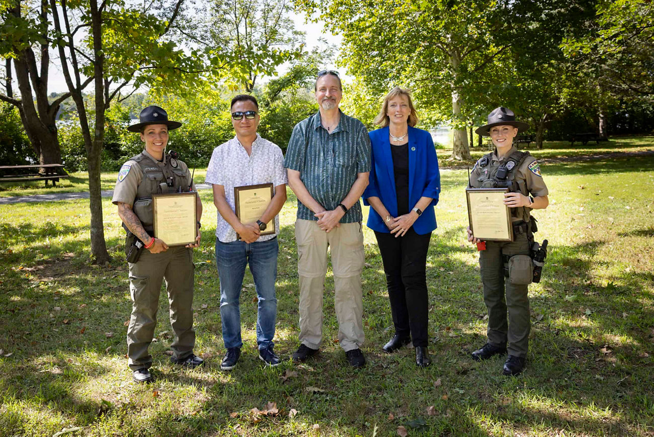 Five individuals stand outdoors on a grassy area surrounded by trees. Two uniformed personnel are positioned at the far left and far right, each holding a framed certificate. Three other individuals stand between them, dressed in casual or professional clothing. The group is posed side by side, facing the camera, with sunlight filtering through the trees in the background.