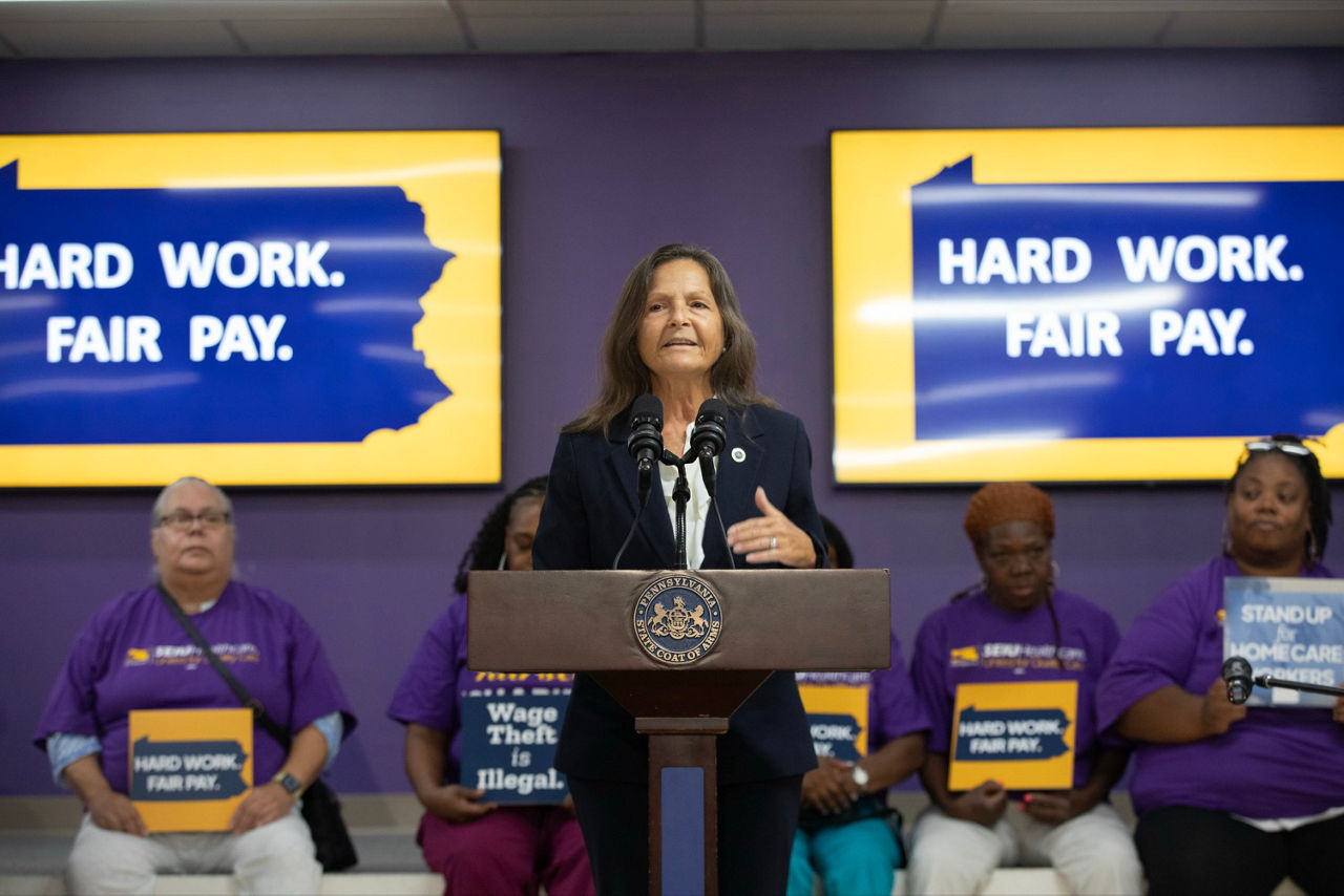 L&I Secretary Nancy A. Walker speaks at a podium before workers in purple shirts, with signs reading “HARD WORK. FAIR PAY.” at a home care worker event.