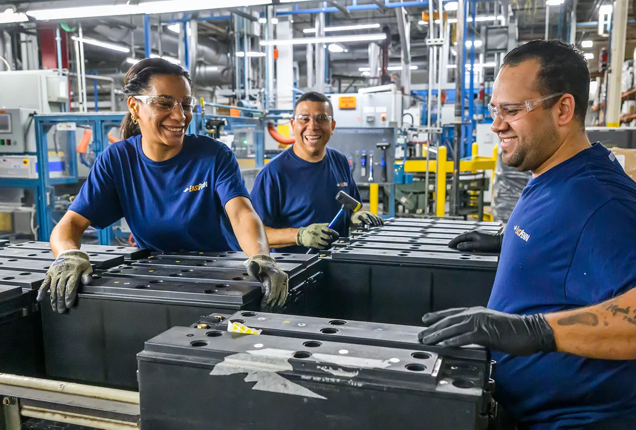Three manufacturing employees wearing shirts with an East Penn company logo smile as they work on a modern manufacturing line.