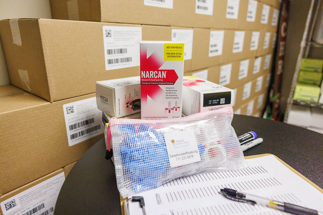 A close-up of an overdose prevention kit assembled by Sara's House of Hope, containing state-supplied Narcan and other harm reduction resources, displayed on a table with additional Narcan supplies in the background.