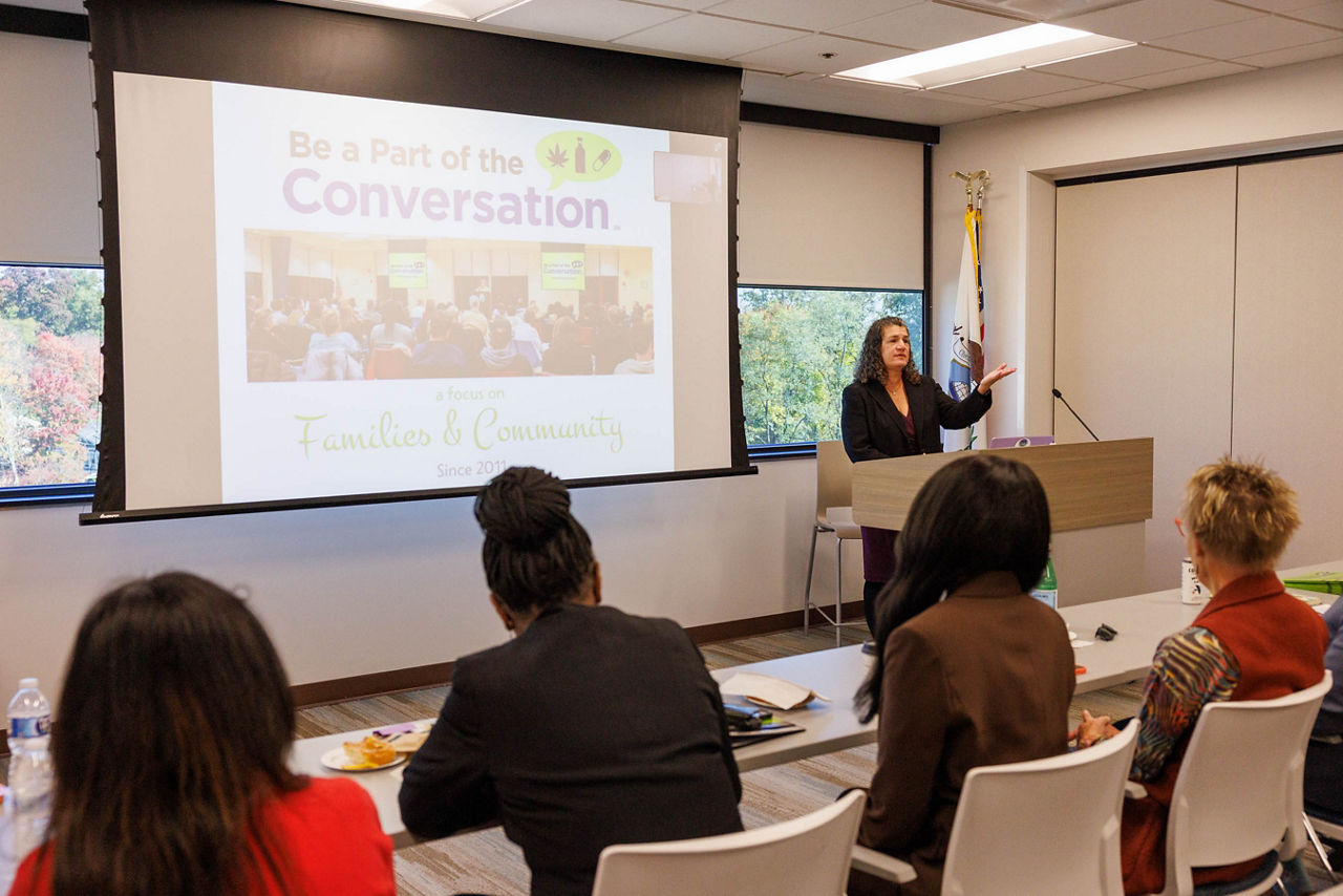 Kathleen Collier, Prevention Program Manager at Chester County Drug & Alcohol Services, speaks at a podium in front of a presentation screen reading “Be a Part of the Conversation: A Focus on Families & Community,” as Pennsylvania Department of Drug and Alcohol Programs Secretary Dr. Latika Davis-Jones looks on from the audience.