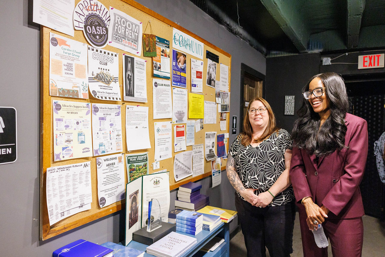 Secretary Latika Davis-Jones and OASIS Community Recovery Club President Danielle Houtz stand at the Club's bulletin board during the Secretary's visit to the Club