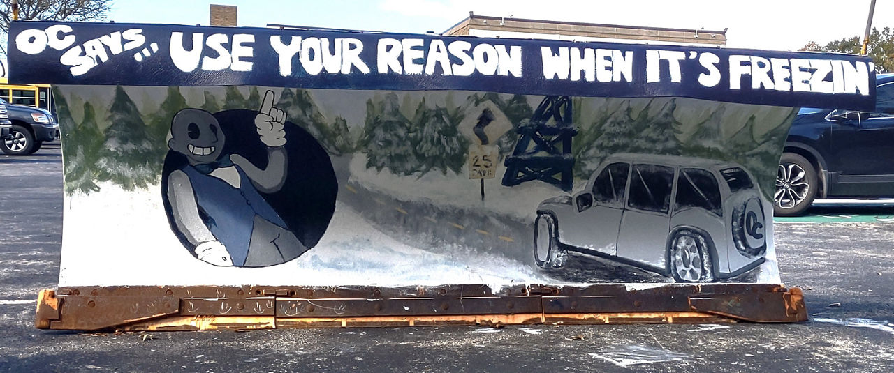A plow painted by students at Oil City High School depicting a car traveling on winter roads with a character pointing to the safety slogan "Use Your Reason When It's Freezin."