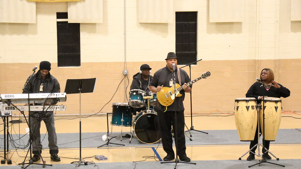 A band consisting of four musicians playing the guitar, keyboard, drums and bongo inside a prison gym.