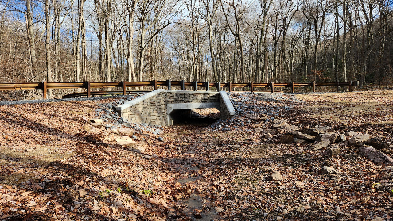 A small stone‑faced box culvert sits beneath a gravel and leaf‑covered roadway area in a forest of tall, leafless trees. The culvert opening is surrounded by rocks and slopes gently downward toward a dry, leaf‑covered streambed. A wooden guardrail runs along the road above the culvert, and the forest extends into the background.