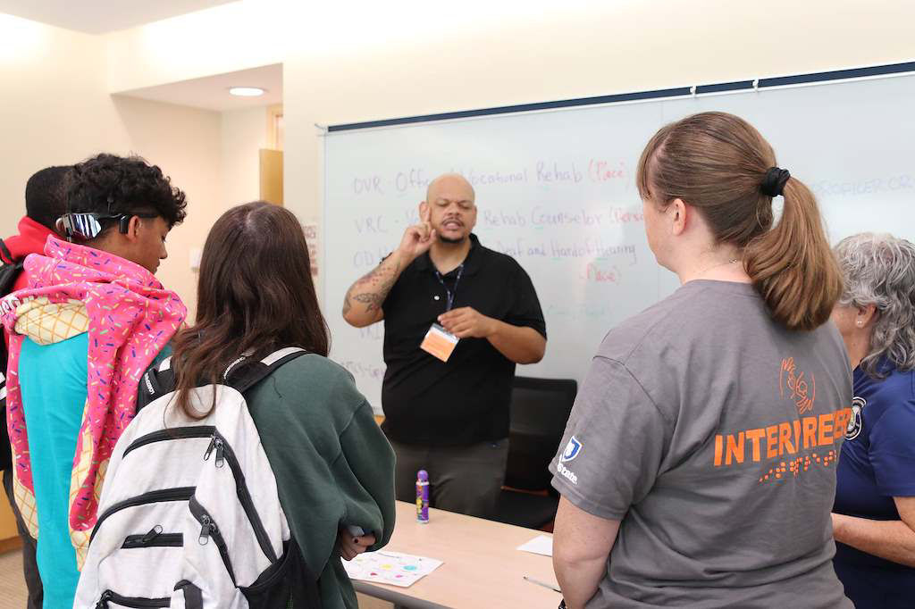 A diverse group of individuals gathered around a ASL interpreter while signing. 