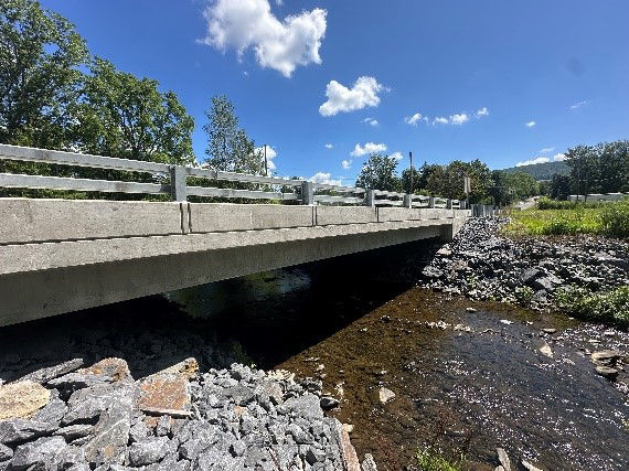 A concrete bridge over a small creek with stones in the foreground and grass and trees in the background.