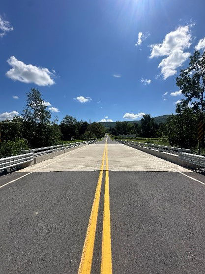 A two-lane roadway divided by a double yellow line with trees, blue skies and white clouds in the background.