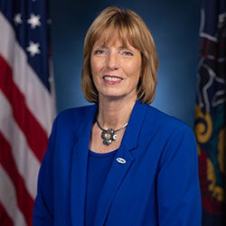 Official Portrait of DCNR Secretary Cindy Adams Dunn wearing a blue suit in front of American and Pennsylvania flags