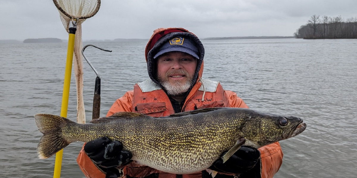 Field Biologist with coat hood up on a boat holding Walleye 