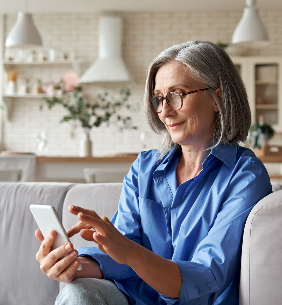 A woman with trendy, bob-style gray hair and red glasses looks at her phone while sitting on a gray couch.