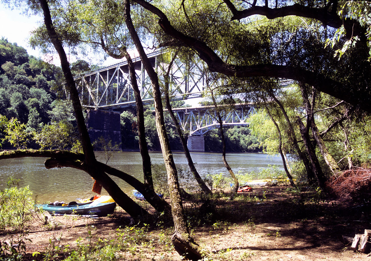 A kayak sitting along the river near an iron bridge over the river