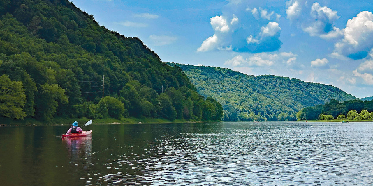Wide shot of a kayaker wearing a life jacket while paddling the Allegheny River with a beautiful scenic mountain in the background.
