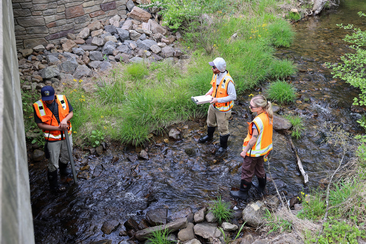 Three people wearing orange vests and taking notes while standing on a stream examining a overpass.