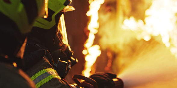 Two firefighters hold up a hose, pointed toward a blaze.