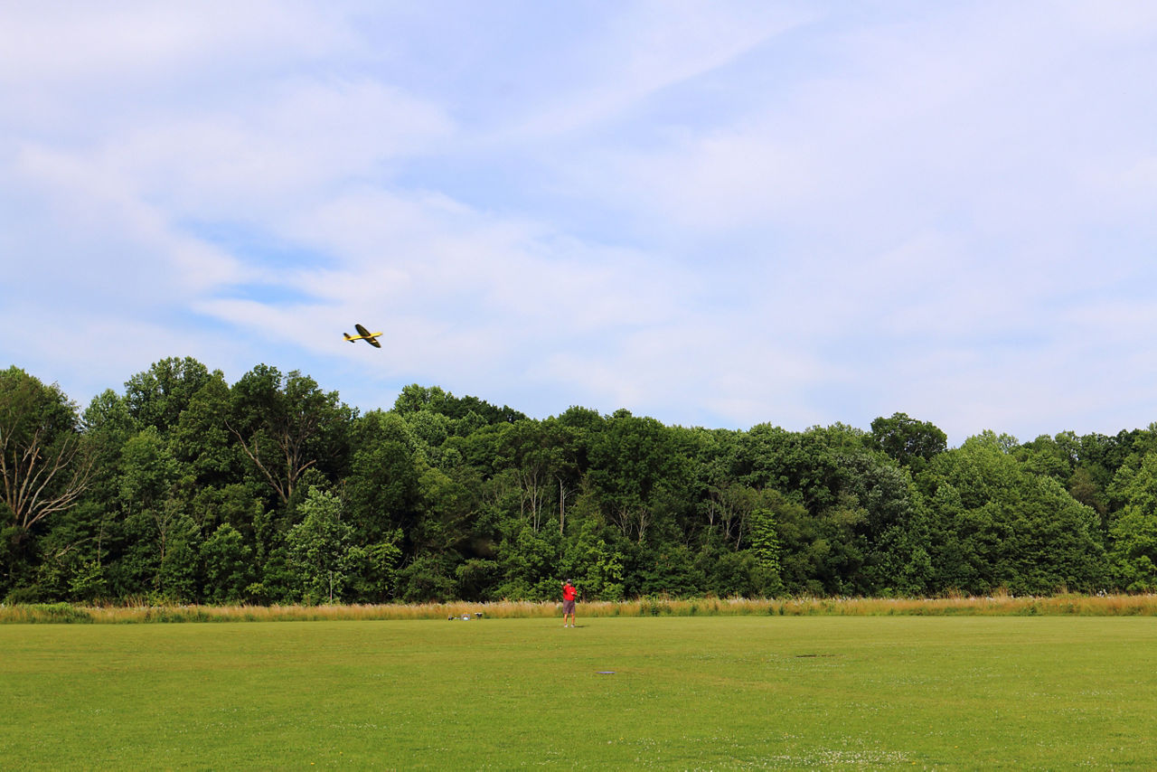 A man flying a red, remote-controlled model airplane over a grassy are with forest in the background