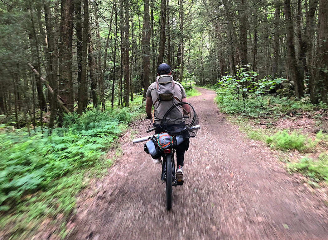 Man with fishing gear and a net biking on a dirt trail in a forest.