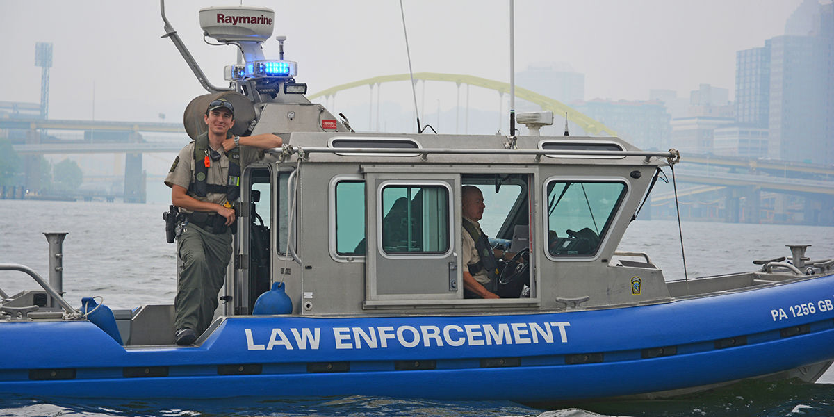 WCO Matt Smith wearing a life jacket and uniform while standing on the side of a PFBC Law Enforcement boat driven by another fellow WCO with the Pittsburgh skyline in the backdrop