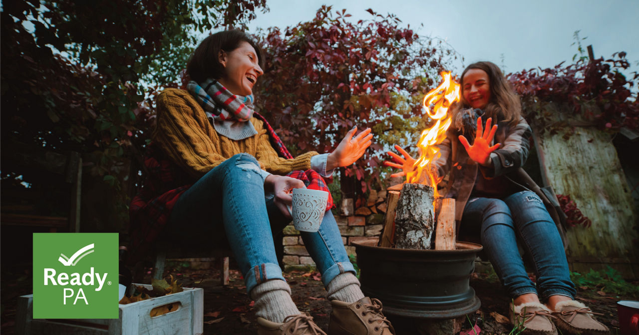 Two women enjoy a bonfire in front of fall foliage.