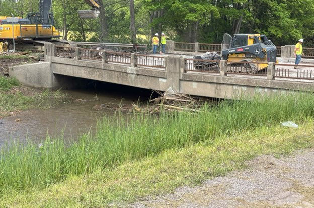 Roadway maintenance crews use various pieces of equipment to demolish a bridge over a creek.