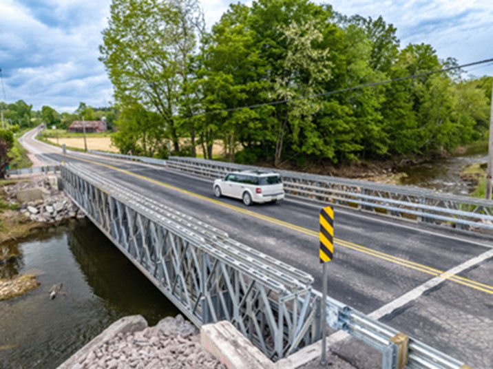 A car drives on the reconstructed Boynton Bridge over Piney Creek. The background is lined with green trees.