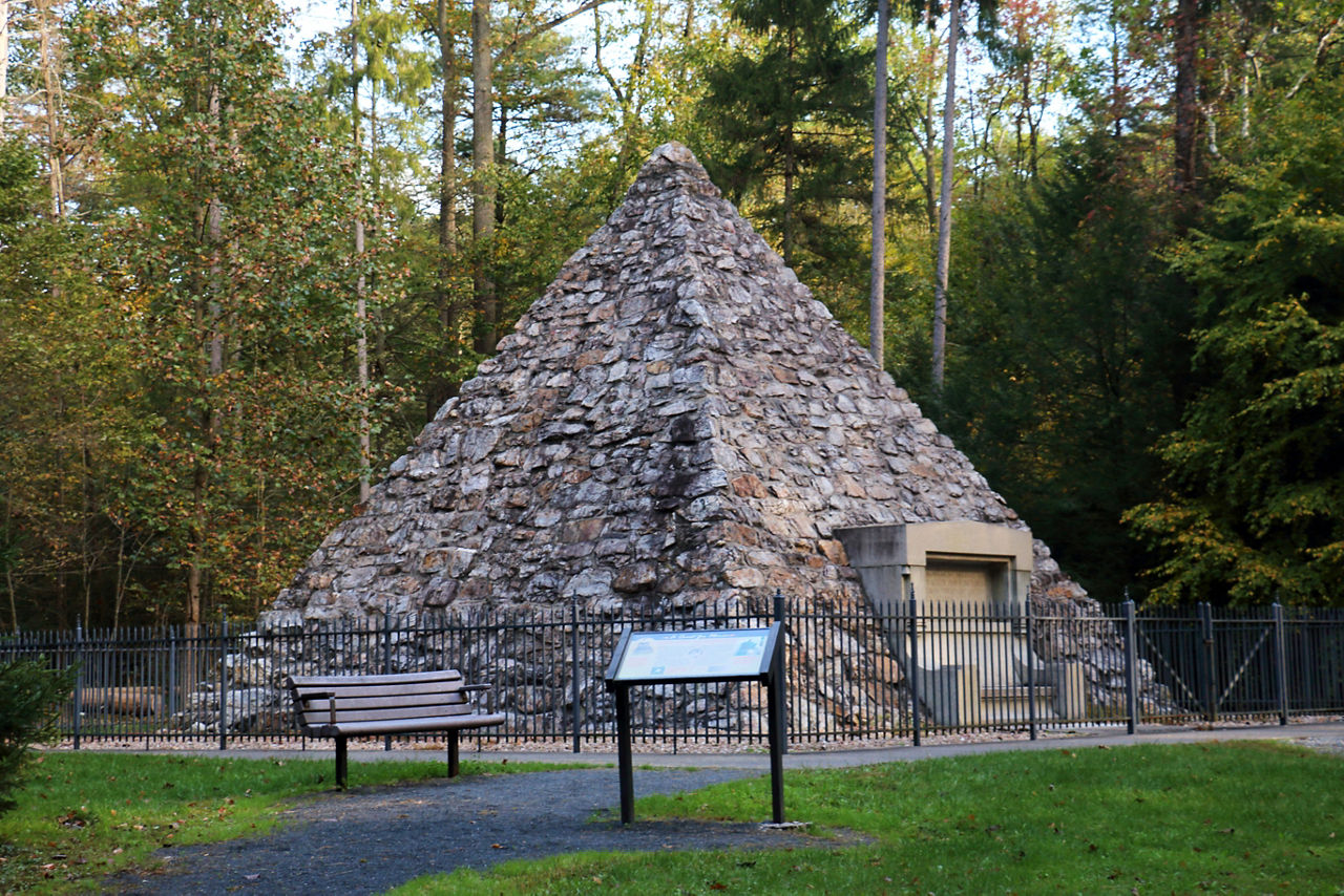 A fenced in historic pyramid-shaped monument with a bench and information panel outside the fence