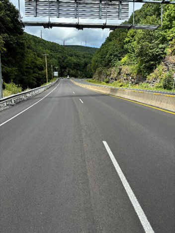 A tree-lined, four-lane roadway divided by a concrete median barrier with a large highway directional sign affixed to a metal pole hanging over the roadway. 
