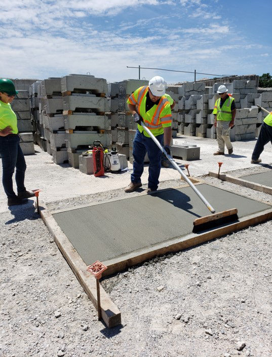 An individual in a hard hat and safety vest using a broom to finish a concrete pavement pad.