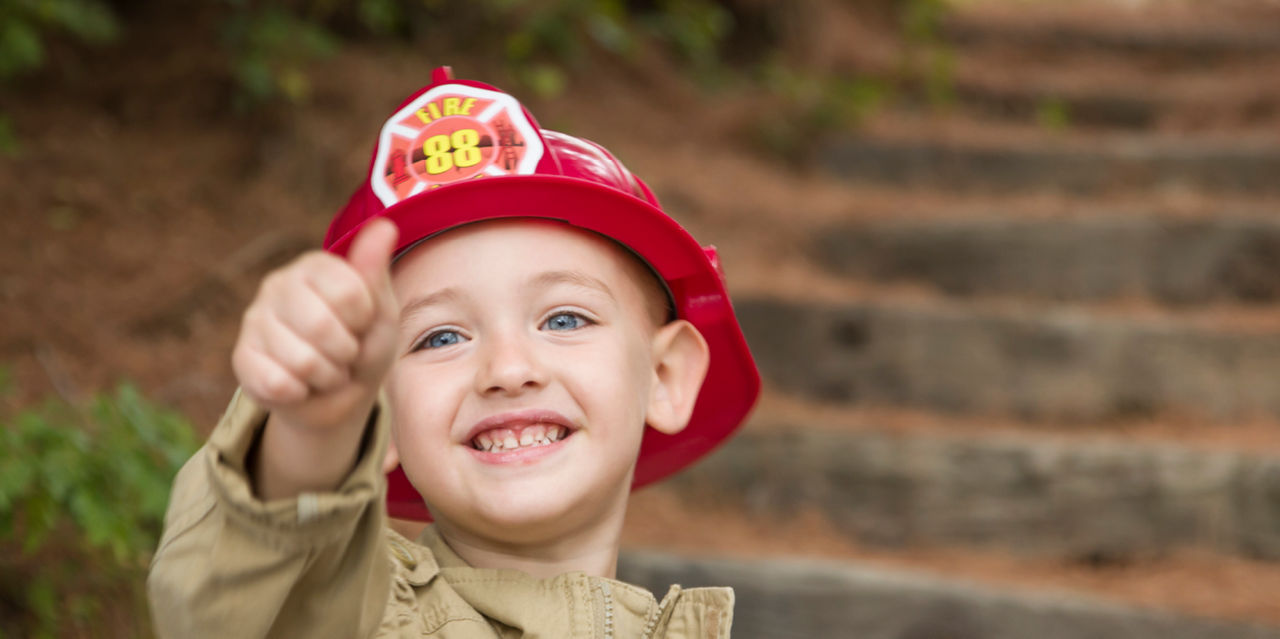 A young boy in a play firefighter helmet gives a thumbs up.