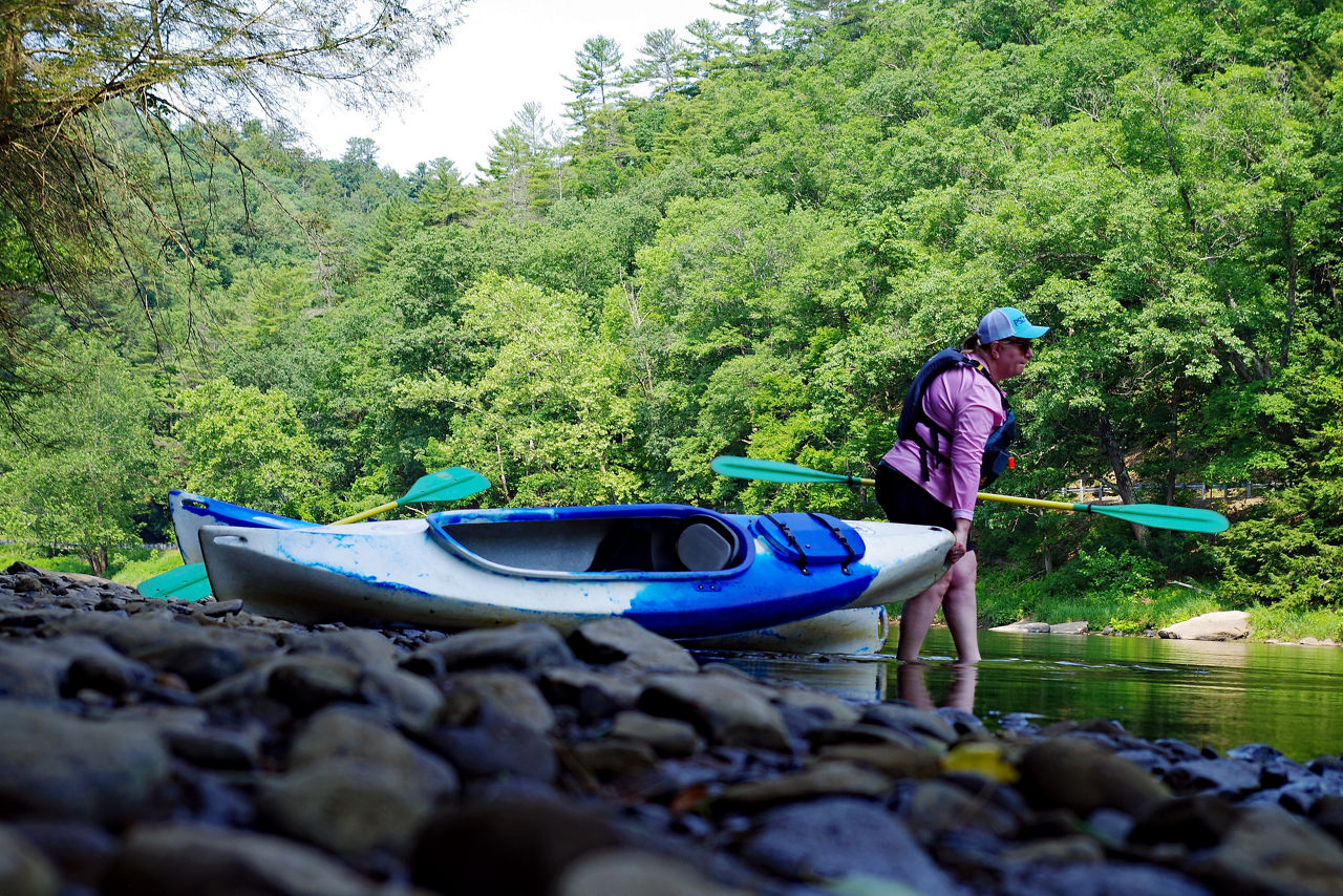 A woman wearing a life jacket pulls her kayak into the Clarion River along a rocky shoreline.