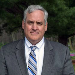 Ken Kane smiles outdoors in front of tree wearing a suit and tie