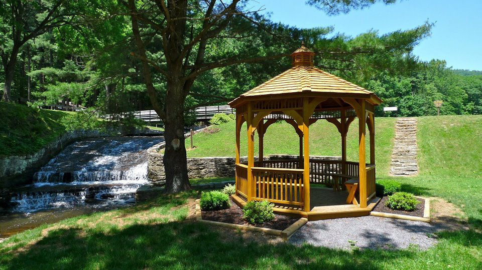 A wooden gazebo surrounded by grass and a manmade waterfall on a sunny day