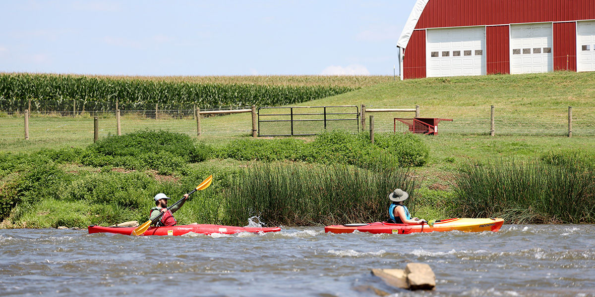 Two people wearing life jackets paddling in kayaks on the Conestoga River looking at a barn.