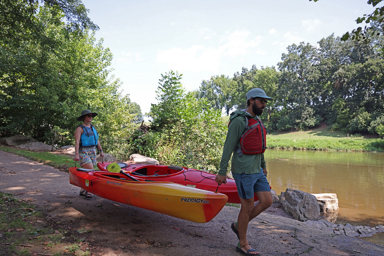 Two people wearing life jackets carrying kayaks down a boat launch along the Conestoga River.