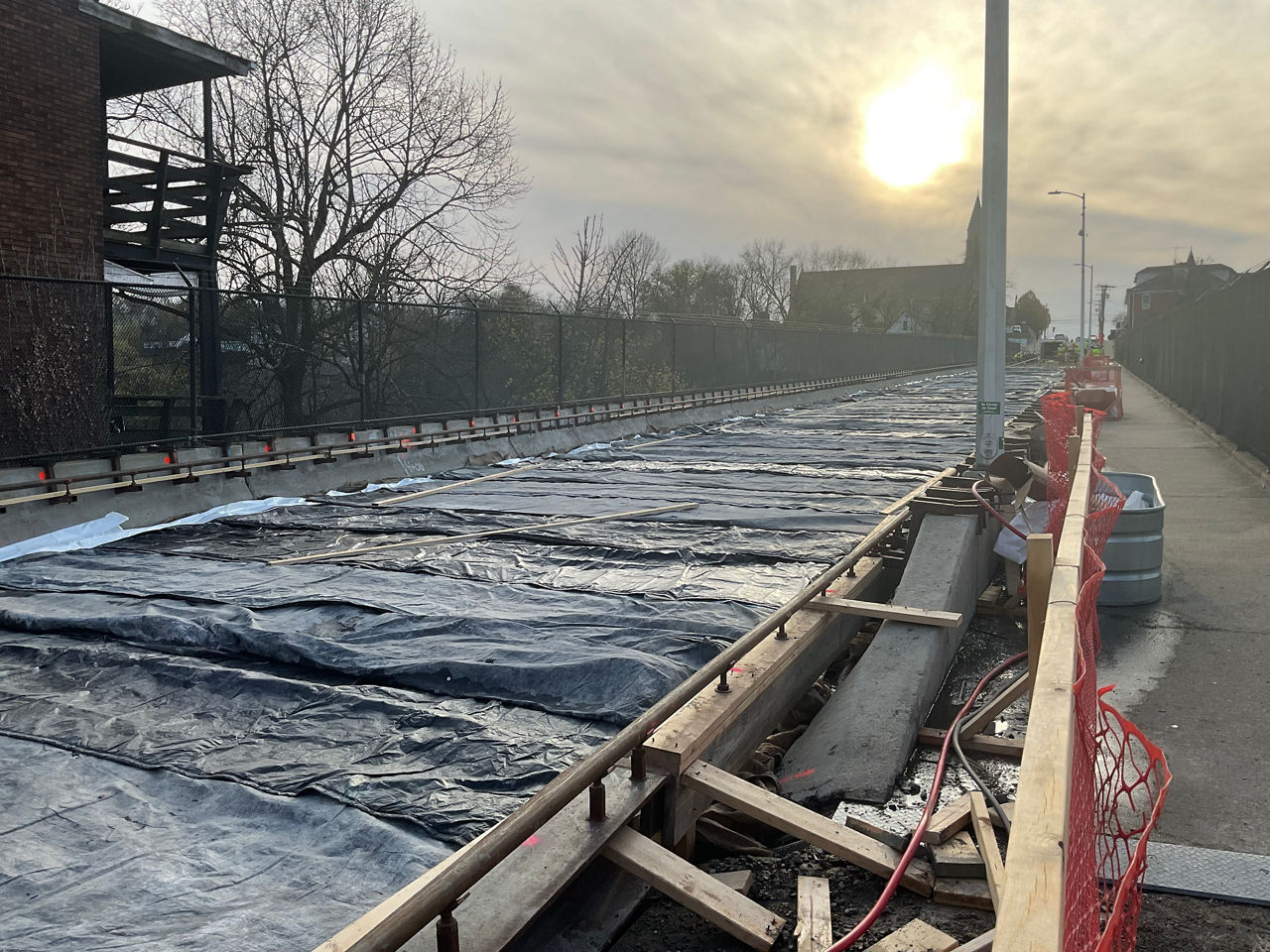 A bridge under construction is covered with burlap. Orange construction fence is in the foreground while sun is peeking out through the clouds is in the background.