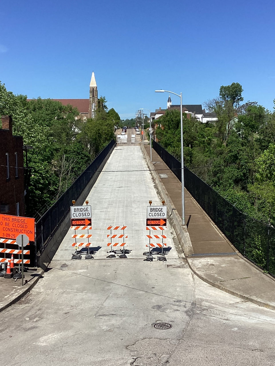 Road closure signs sit at the end of a concrete bridge lined with green trees on either side. A church steeple is visible in the background.
