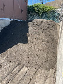 A large mound of soil sits next to a building with dirt tracks in the foreground and green shrubbery in the background.