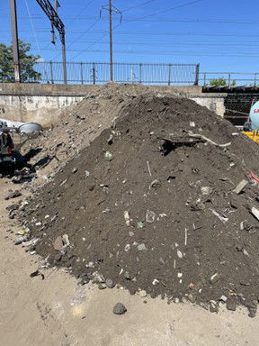 Two large mounds of dirt and debris since on concrete pavement. A large metal fence and electric lines are in the background.  