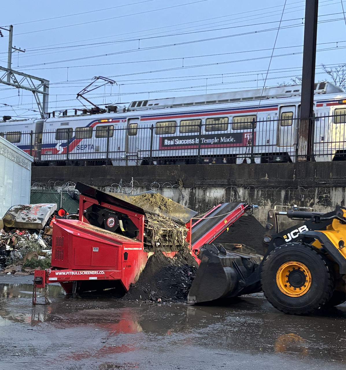 A yellow backhoe loads materials into a large red piece of equipment mounted on a trailer chassis with a conveyor belt on one end and a trailer hookup on the other end.